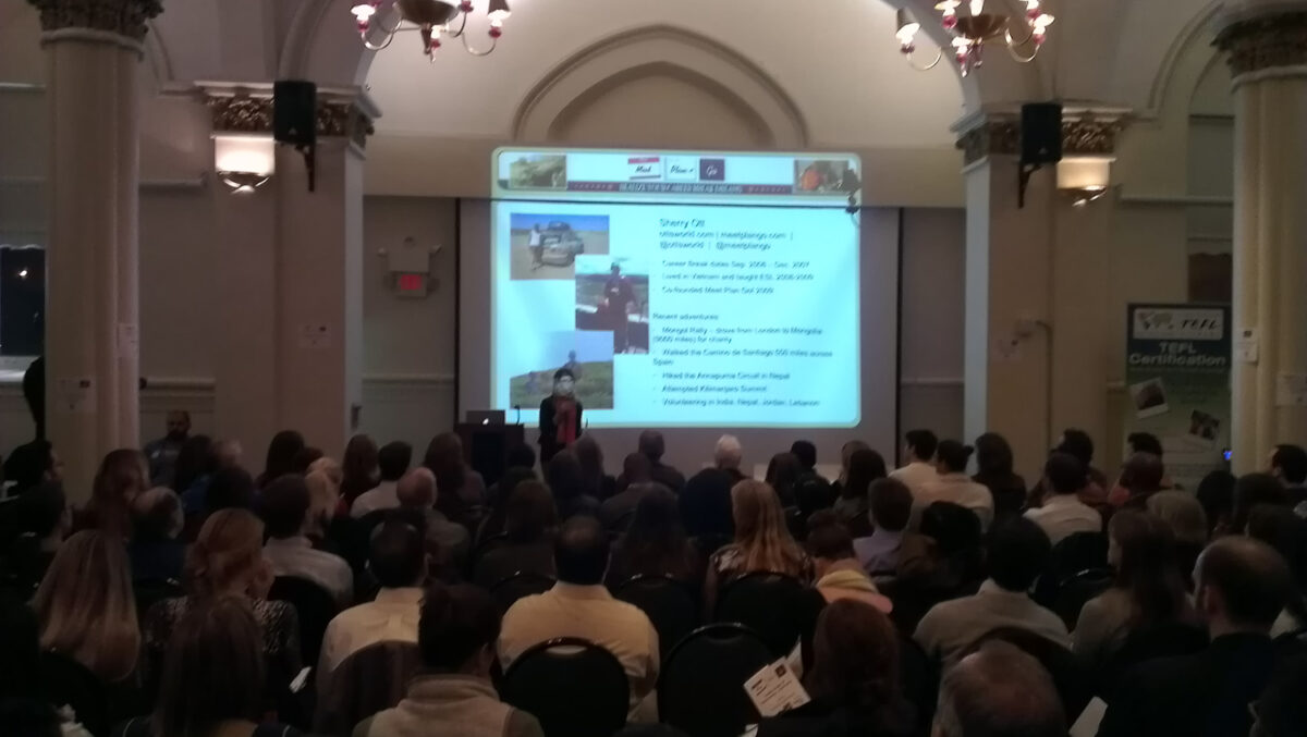 Speaker presenting at a conference with an audience in a historic hall with chandeliers.