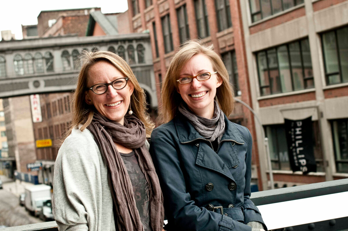 Two smiling women with glasses and scarves standing outdoors in an urban setting with historic and modern buildings.