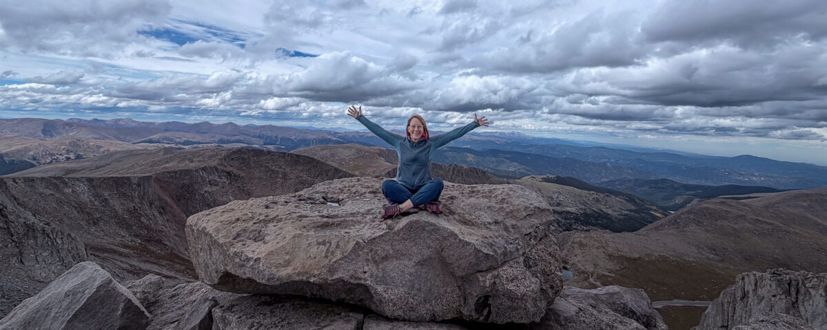 Woman sitting cross-legged on a large rock at a mountain summit with arms raised, overlooking rugged mountain landscape under cloudy sky.