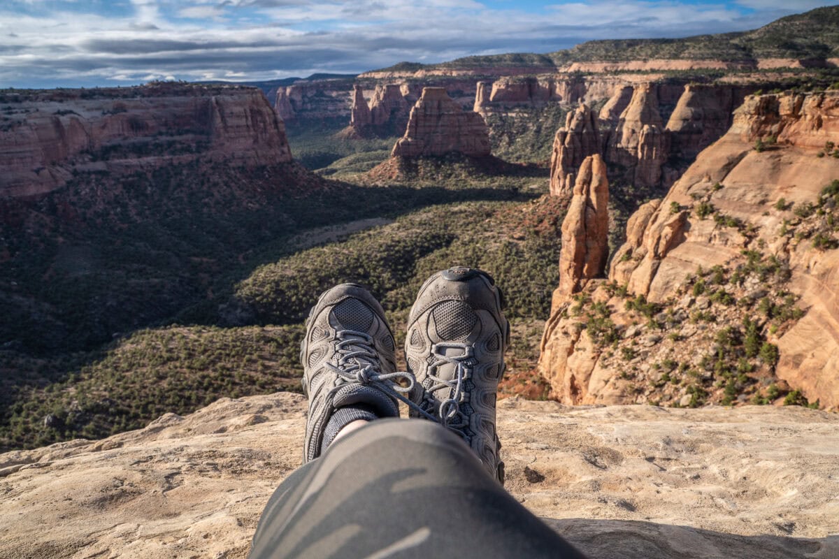 View from the edge of the Grand Canyon showing rugged cliffs and deep valleys, with a hiker's legs and boots in the foreground.