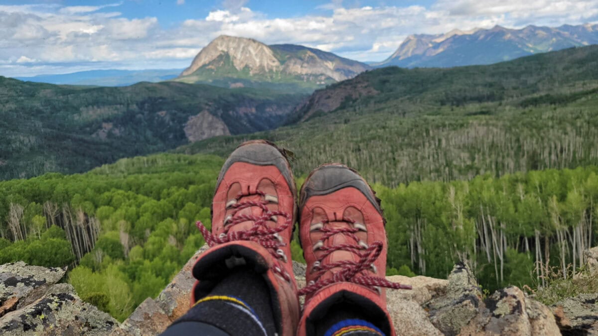 Hiker's red boots resting on a rocky ledge overlooking lush green valleys and towering mountain peaks in the Rocky Mountains.