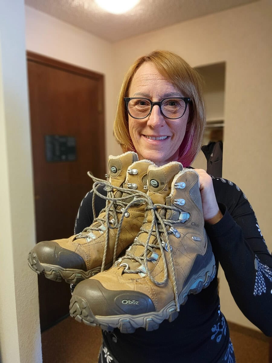 Woman holding a pair of tan hiking boots, smiling, celebrating OttsWorld's 20th anniversary with a giveaway.